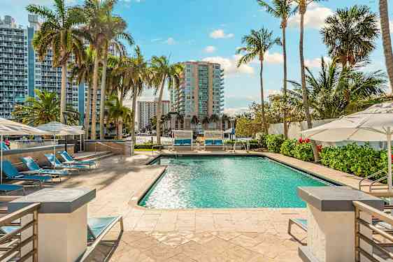 En swimmingpool omgivet af liggestole og parasoller, med høje bygninger og palmer i baggrunden. (AI-genereret tekst)