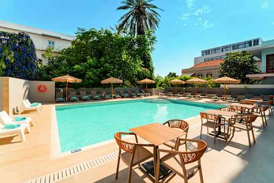 En udendørs pool med liggestole og parasoller på siden samt borde og stole i forgrunden. (AI-genereret tekst)