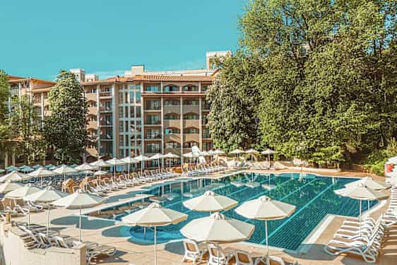 Poolområde med liggestole og parasoller, i baggrunden ses en hotelbygning og grønne træer. (AI-genereret tekst)