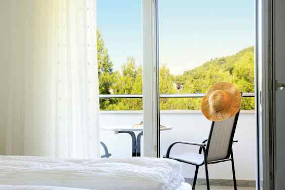 Et hotelværelse med udsigt til en balkon, hvor en stol med en hat og et bord står, samt frodige træer i baggrunden. (AI-genereret tekst)