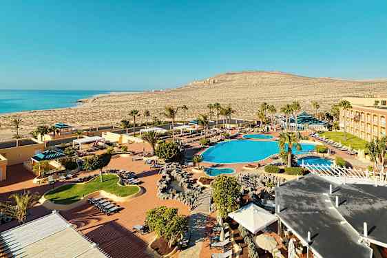 Billedet viser et feriested ved en kyst med poolområder, liggestole og palmer. I baggrunden ses havet og det omgivende tørre landskab. (AI-genereret tekst)