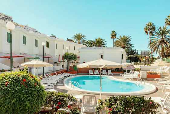 Udendørs swimmingpool med liggestole og parasoller ved hvide bygninger omgivet af palmer. (AI-genereret tekst)