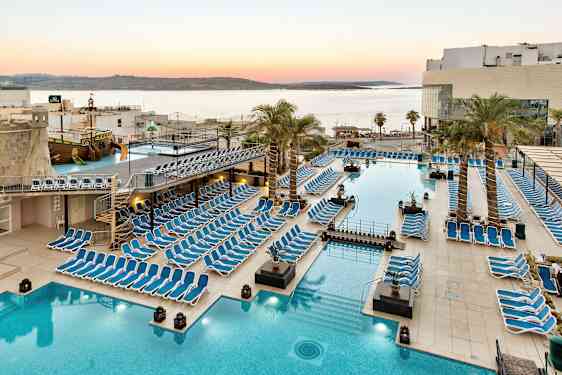 Stort poolområde med liggestole, palmer og udsigt over havet ved solnedgang. (AI-genereret tekst)