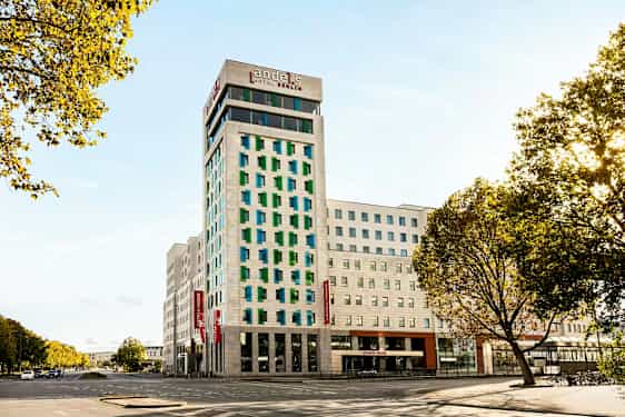En moderne hotelbygning med hvid facade og farvede vinduer, omgivet af veje og træer. (AI-genereret tekst)