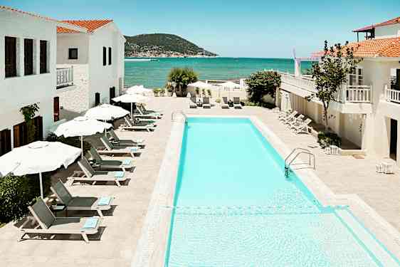 Billede af en udendørs swimmingpool med liggestole og parasoller, omgivet af hvide bygninger med udsigt over havet. (AI-genereret tekst)