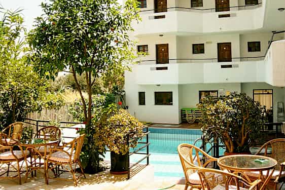 En udendørs pool omgivet af beplantning og siddepladser samt en hvid bygning med altaner i baggrunden. (AI-genereret tekst)