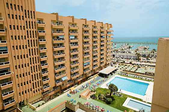 En høj bygning med altaner og en pool foran samt en marina i baggrunden. (AI-genereret tekst)