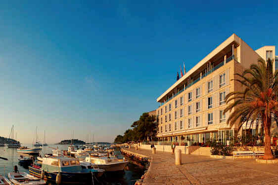 Billedet viser en kystlinje med små både fortøjet ved kajen og en moderne bygning omgivet af palmer i baggrunden. (AI-genereret tekst)