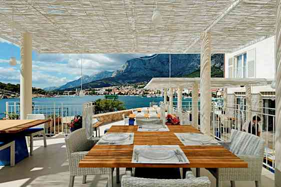 En terrasse med udsigt over bjerge og hav, flere borde er dækket til gæster. (AI-genereret tekst)