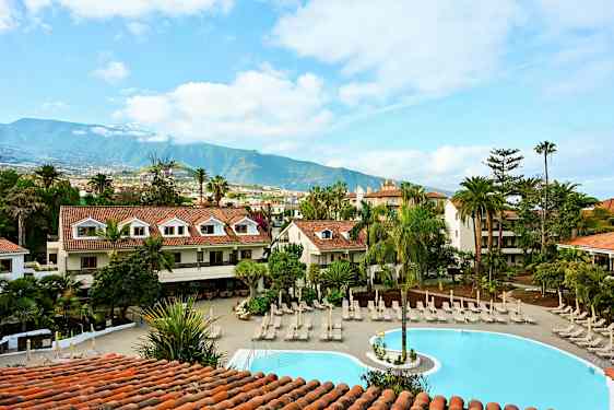 Et hotelområde med en pool omgivet af liggestole og grønne områder med bjerge i baggrunden. (AI-genereret tekst)