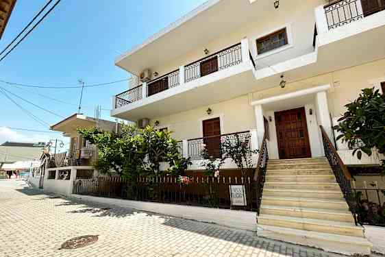 En hvid bygning med altaner og en trappeopgang ses fra en stenlagt gade. (AI-genereret tekst)