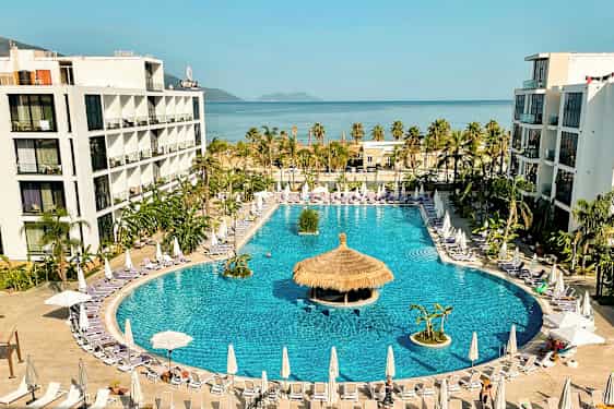 Hotelkompleks med stor swimmingpool omgivet af liggestole og parasoller, med havet i baggrunden. (AI-genereret tekst)