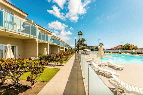 Billede af et hotelområde med pool, liggestole og grønne omgivelser. Bygningen har altaner. (AI-genereret tekst)