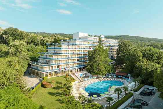 Billede af et hotel med flere etager og altaner, omgivet af frodigt grønt. En pool med liggestole ligger i forgrunden. (AI-genereret tekst)