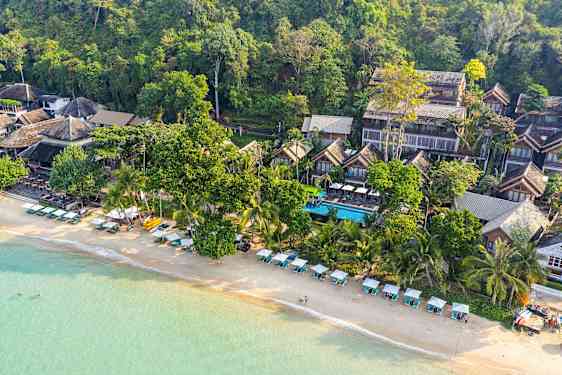 Luftfoto af en strand med liggestole og parasoller samt bygninger omkranset af grønne områder. (AI-genereret tekst)