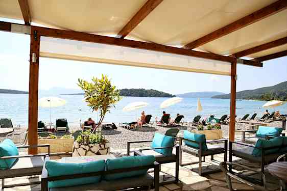 Udsigt fra et skyggefuldt siddeområde mod en strand med liggestole og parasoller ved en rolig havvig. (AI-genereret tekst)