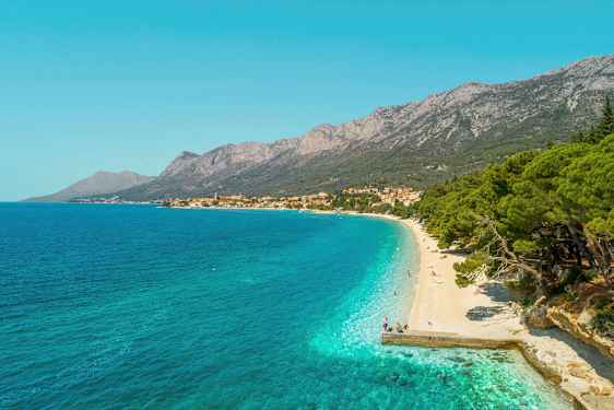 Udsigt over en stenstrand med turkis vand og frodige træer. Bjerge og en landsby ses i baggrunden. (AI-genereret tekst)