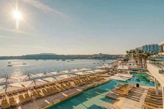 Udsigt over et solrigt poolområde med solsenge og parasoller, med havet og både i baggrunden. (AI-genereret tekst)