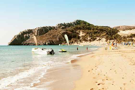 En strand med klart vand, solsenge og parasoller, samt en motorbåd og en bjergrig baggrund. (AI-genereret tekst)