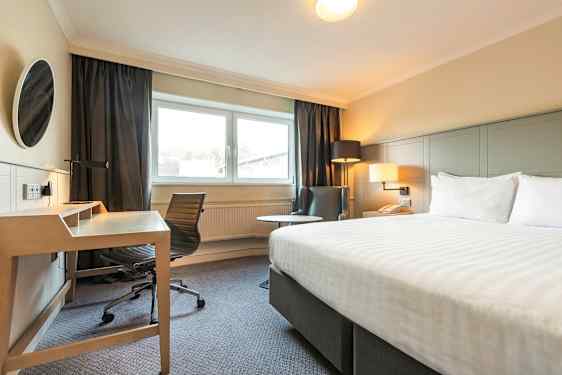 Billede af et hotelværelse med en seng, skrivebord og stol, samt en lænestol ved siden af et vindue. (AI-genereret tekst)