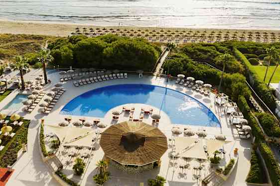 Udsigt over et poolområde med liggestole og parasoller, omgivet af grønt og tæt på stranden. (AI-genereret tekst)