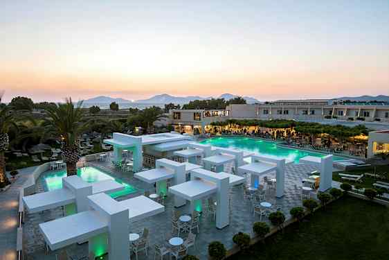 Moderne hotelområde med pool og aftenbelysning, omgivet af grønne områder og palmer med bjerge i baggrunden. (AI-genereret tekst)