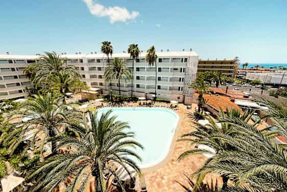 En udendørs pool omgivet af liggestole og palmer. I baggrunden ses et hotel med hvid facade. (AI-genereret tekst)