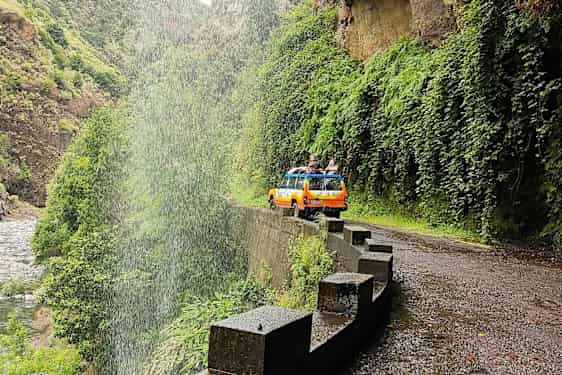 En gul-orange jeep på en vej ved et vandfald i en grøn dal. (AI-genereret tekst)