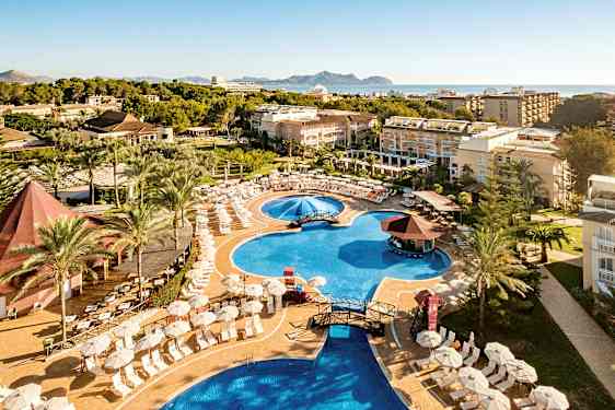 Hotellets poolområde med liggestole, parasoller og omkringliggende bygninger, med havet i baggrunden. (AI-genereret tekst)