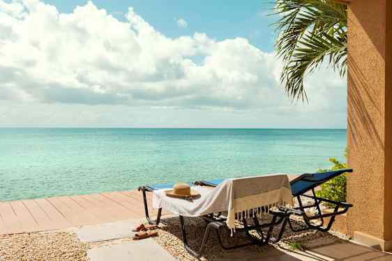 To liggestole med et håndklæde og en hat står ved siden af et lille terrassebord nær havet. En palmegren ses til højre. (AI-genereret tekst)