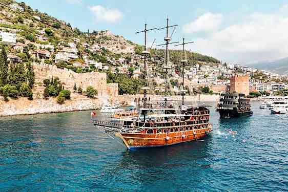 En træbåd med flere master sejler i en blåbugt nær en by på en skråning og en gammel fæstning. (AI-genereret tekst)