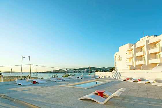 Udendørs poolområde med moderne liggestole og udsigt over havet samt hvide bygninger i baggrunden. (AI-genereret tekst)