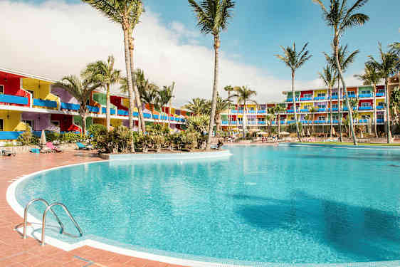 En stor pool omgivet af palmer, med farverige hotelbygninger i baggrunden. (AI-genereret tekst)