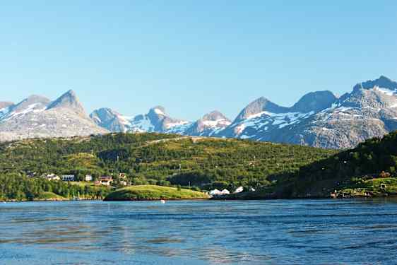 En udsigt over et bjerglandskab med snedækkede toppe, grønt landskab og huse ved en vandflade. (AI-genereret tekst)