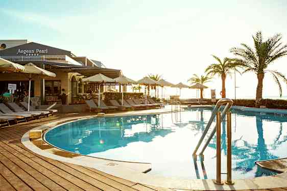 Udendørs pool med liggestole og parasoller omkring. Bygning med skiltet 'Aegean Pearl' ses i baggrunden. (AI-genereret tekst)