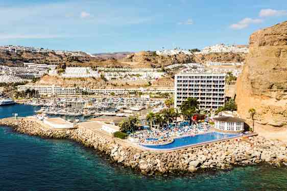 Billede af en kystby med marina, bygninger på skråning og hotel med pool. (AI-genereret tekst)