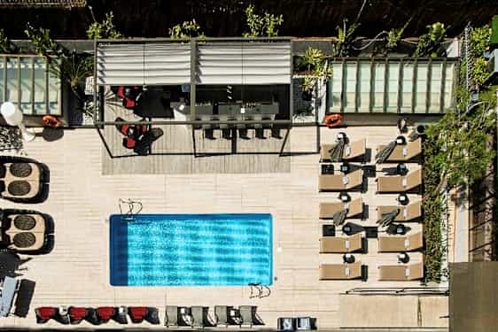 Fugleperspektiv af en udendørs swimmingpool med omkringliggende liggestole, parasoller og en overdækket siddeplads. (AI-genereret tekst)