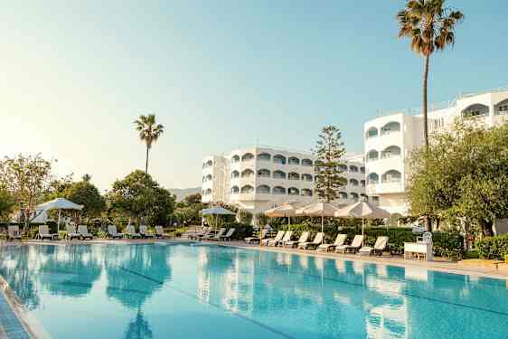 Hotelbygning med pool, liggestole og parasoller i forgrunden. (AI-genereret tekst)