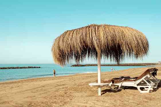 Billede af en sandstrand med to liggestole under en halmparasol samt personer, der går langs vandet. (AI-genereret tekst)