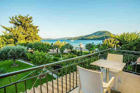 En balkon med bord og stole, udsigt over en grøn have, havet og bjerge i baggrunden. (AI-genereret tekst)