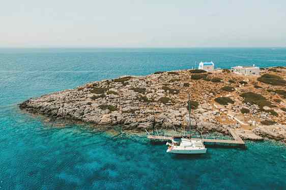 En kyst med klipper, turkisvand og en hvid sejlbåd ved en bro tæt på små bygninger. (AI-genereret tekst)