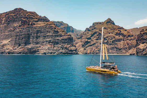 En katamaran sejler tæt på høje klipper, med klart blåt vand i forgrunden. (AI-genereret tekst)