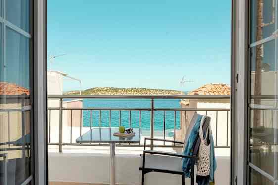 Udsigt fra et værelse gennem døre mod en balkon med bord, stol og tøj, samt hav og landskab i baggrunden. (AI-genereret tekst)