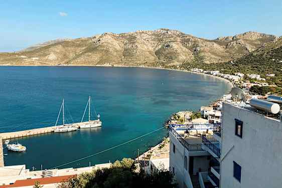 Udsigt over en kyst med små bygninger og bjerge i baggrunden. Både ligger fortøjet ved molen i det klare vand. (AI-genereret tekst)
