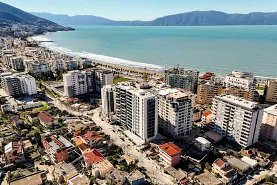 Billedet viser en by med højhuse og boliger tæt på en kystlinje med sandstrand og palmer. (AI-genereret tekst)