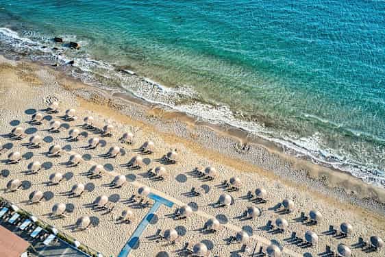 Luftfoto af en sandstrand med liggestole og parasoller, med kystlinjen og blåt hav i baggrunden. (AI-genereret tekst)