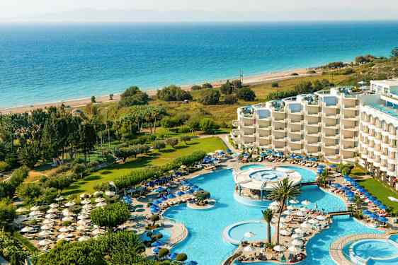 Udsigt over et resort med poolområder, liggestole, grønne områder og havet i baggrunden. (AI-genereret tekst)