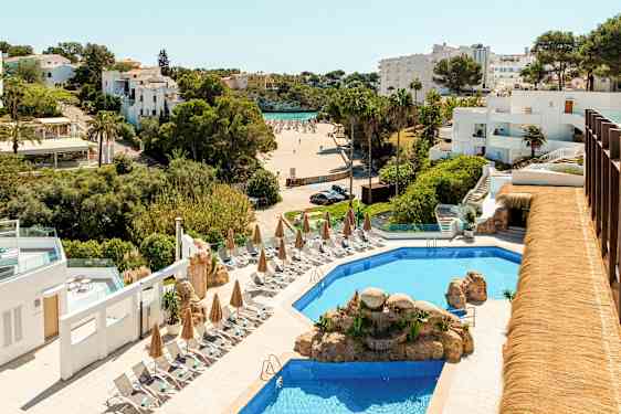 Hotelområde med en pool omgivet af liggestole og parasoller, i baggrunden ses en strand og hvidmalede bygninger. (AI-genereret tekst)