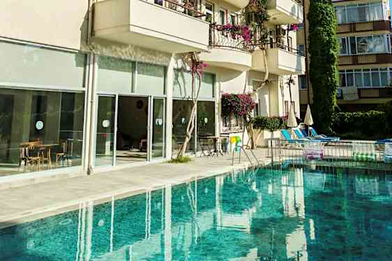 En pool foran en bygning med altaner og store glasvinduer. Billederne viser også stole og borde samt parasoller ved poolområdet. (AI-genereret tekst)