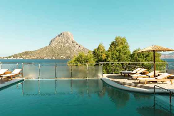 En udendørs pool med liggestole og parasol, beliggende tæt på havet med en bakkelignende høj i baggrunden. (AI-genereret tekst)
